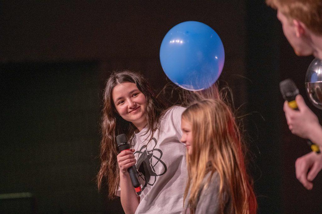 Student wearing an Engineering and Physics Club white shirt, with long brown hair smiling at a young girl with long blonde hair in front of her. The student is holding an inflated blue balloon over the young girls head and her hair floats up toward the balloon.